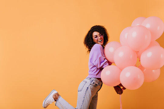 Good-humoured Birthday Girl In Jeans Posing On Orange Background. Enchanting Black Lady Dancing With Party Balloons.