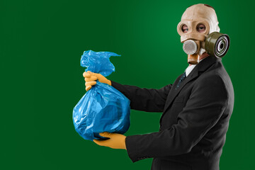 Portrait of a man wearing a business suit and an old gas mask holding a blue garbage bag, isolated on green background.