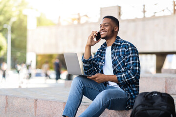 Black man using laptop, sitting in the city with smartphone