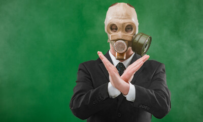 Portrait of a man wearing a business suit and an old gas mask with his hands x crossed, isolated on green foggy background.