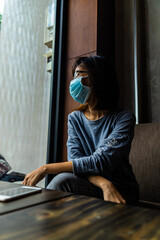 Obraz premium Quarantine self-isolation. Sadness young asian woman in a medical mask who sit on sofa and looks out the window through the window.