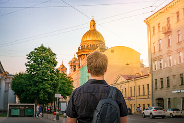 A young blond man stands on the street of Saint Petersburg against the background of the dome of St. Isaac's Cathedral in the summer, photo from the back