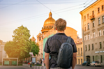 A young blond man stands on the street of Saint Petersburg against the background of the dome of St. Isaac's Cathedral in the summer, photo from the back