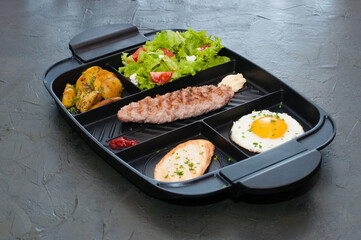 Breakfast, cutlet or steak, tomato salad, fried potatoes, croutons and fried egg on black plate. Restaurant lunch, content for the site