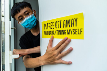 Young asian man setting up self quarantine door sign for front of house, because of covid-19...