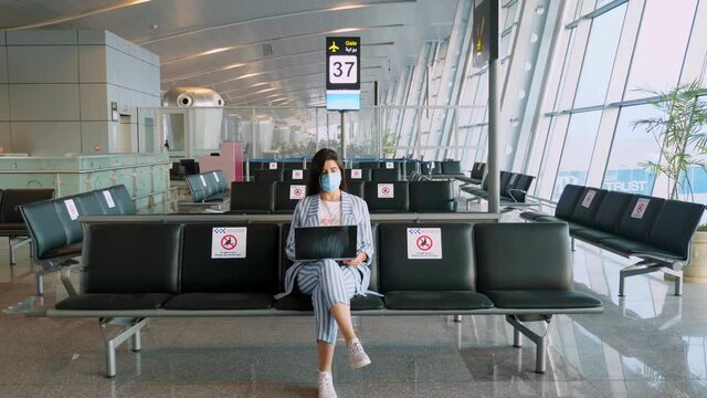 woman, in protective mask, uses laptop, while sitting in empty airport lounge, waiting to board a flight. air travel re-opened after coronavirus outbreak end. Opening borders. travel open.