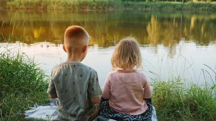 Rear view of a couple of children, a little boy and a girl, are sitting on the grass by a small lake at sunset, talking by throwing pieces of bread into the water at sunset