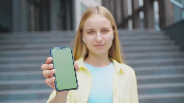 Close up woman shows smartphone with a green screen hold in hands look at camera smile in street display digital chroma key cellphone internet mobile slow motion
