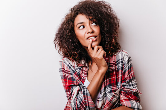 Pensive curly woman looking around and touching her chin. Indoor photo of attractive african female model in checkered shirt isolated on light background.