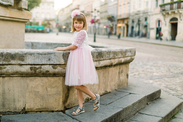 Adorable little blond girl in beautiful pink dress posing to camera at old fountain of ancient European city outdoors. Full length portrait of happy pretty little girl in old city