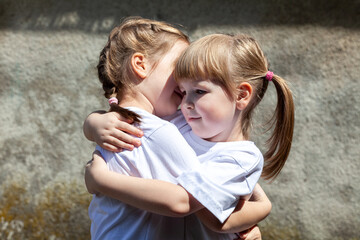 Two little sisters hugging each other smiling. Happy young kids, siblings close together, portrait. White t-shirts, care concept