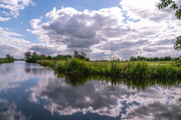 Wide ditch with green vegetation along it. In the ditch, the blue sky with the low-hanging clouds is reflected. Reeuwijk, South Holland, Netherlands, Europe.