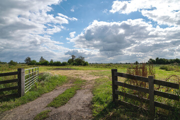Dirt road that ends in grassland with an open entrance gate. Typically Dutch cloudy sky above. Reeuwijk, South Holland, Netherlands, Europe.