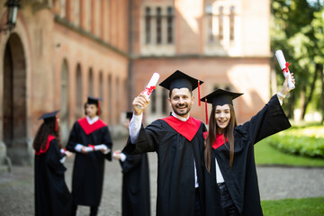 Excited young two graduates in robes standing and showing certificate in front of students