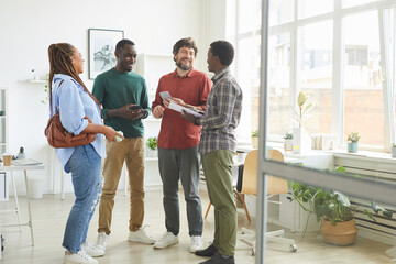 Full length portrait of multi-ethnic group of people dressed in casual wear and smiling cheerfully while discussing work standing in office - Powered by Adobe