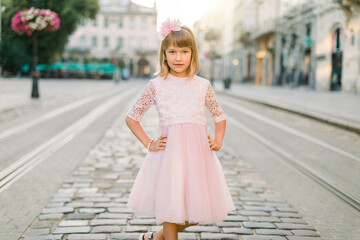 Fototapeta premium Adorable little girl in elegant pink dress, with flower accessories in her blond hair, posing to camera in summer morning in ancient city Lviv, Ukraine. Outdoor city portrait of fashion little girl