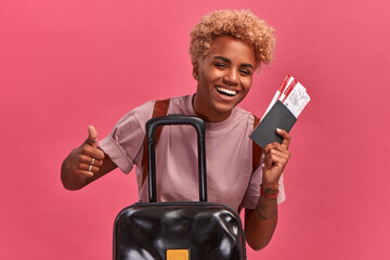 African american woman holds tickets. Joyful tourist received a visa and stands on a background of a pink wall holding tickets and shows with his hand that everything is cool. Summer vacation concept.