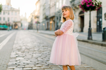 Little girl in princess pink dress walking outdoor and enjoying the view of ancient European city center. Lifestyle portrait of little girl walking in the city