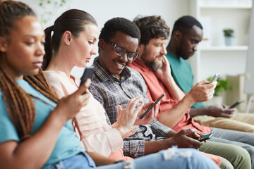 Fototapeta na wymiar Portrait of multi-ethnic group of people sitting in row and using smartphones while waiting for conference, focus on smiling African-American man talking to female colleague beside him, copy space