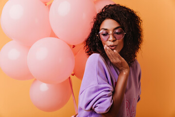 Obraz na płótnie Canvas Ecstatic brunette girl in glasses standing on yellow background with balloons. Indoor shot of lovable african lady posing with surprised face expression.