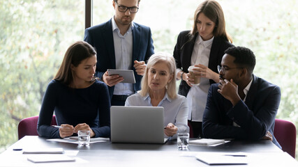 60s woman mentor teach young multi ethnic employees, group of diverse people analyzing online...