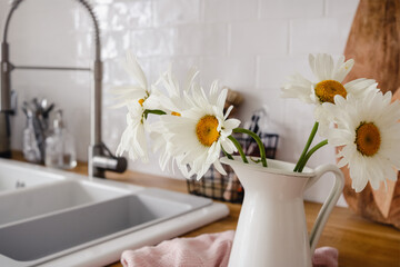 White chamomile flowers in a jug on the kitchen counter with a faucet and a sink in the background
