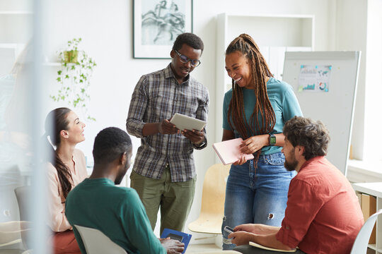 Multi-ethnic group of people sitting in circle while discussing business project in office, focus on smiling African-American woman talking to colleague standing up , copy space
