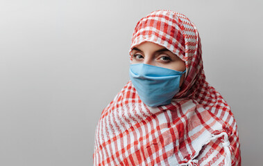 Young muslim woman in keffiyeh and protective medical mask on a gray background looks at the viewer. The concept of quarantine, coronavirus, pandemic COVID-19. Copy space.