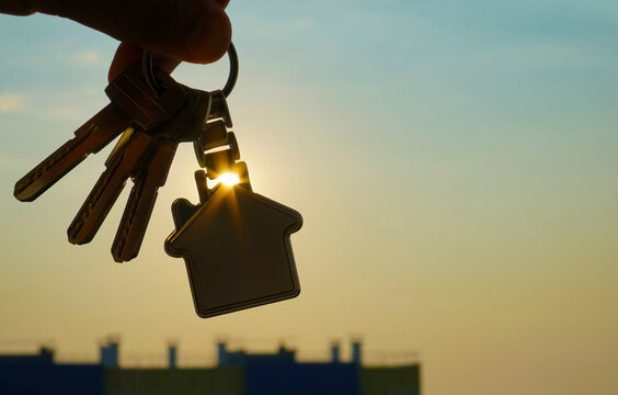 Buying or renting resindental house concept. Man hand holding 3 three silver key with home shape keyring, on sunlight sky cityscapes background. - Powered by Adobe