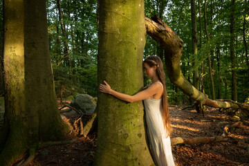 Frau in weißem Kleid umarmt einen Baum im Wald, Naturtherapie und Waldbaden als...