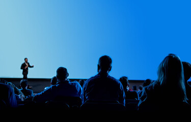 Business or Academic Conference Event. Audience engaged and looking down at a figure on stage. The stage screen is blue. - obrazy, fototapety, plakaty