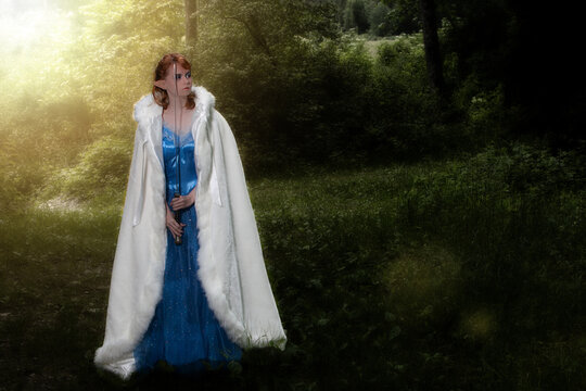 Junge rothaarige Frau mit Elfenohren, blauen Kleid und weißen Umhang hält ein Schwert vors Gesicht.