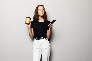 Portrait of a young smiling girl in shirt texting message on mobile phone and holding cup of coffee to go over gray background