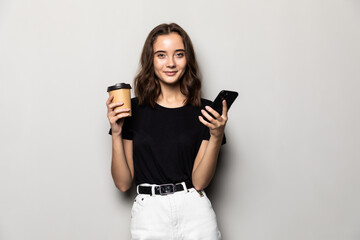 Portrait of a young smiling girl in shirt texting message on mobile phone and holding cup of coffee to go over gray background