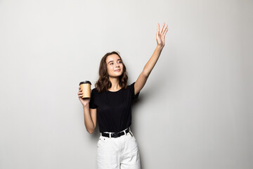 Fototapeta na wymiar Young woman waving hand while walking and holding cup with coffee isolated over gray background