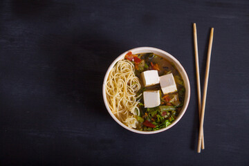 Japanese soup with vegetables, pasta, tofu cheese, chopsticks on a black background. Flat lay.