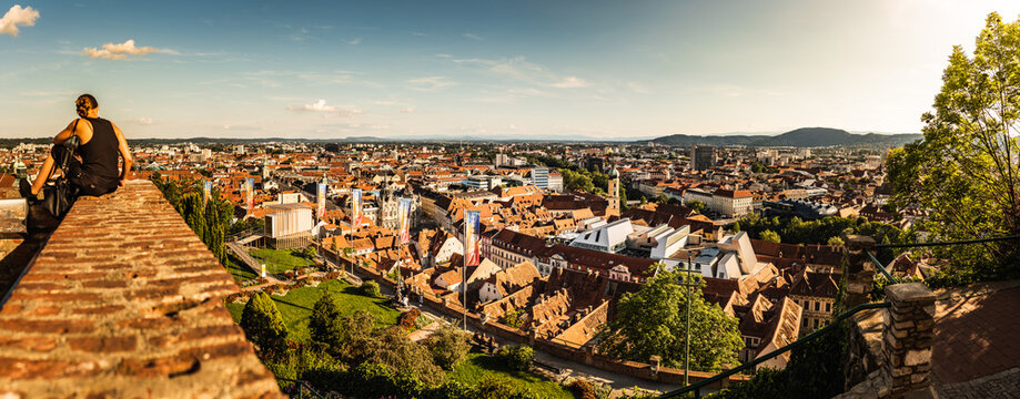 Woman Sitting On Red Wall Against Panorama View From The Top Of Schlossberg Hill Over The City