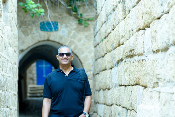 Fototapeta na wymiar Smiling trendy tourist senior with black t-shirt and sunglasses standing by old ancient city street with brick historic stone wall.