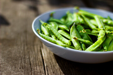 Green pea pods in a white bowl on a wooden background with copy space, vegan food and healthy organic food concept. Harvesting.