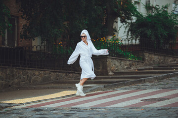 A glamorous lady in a white bathrobe with a white towel on her head and elegant sunglasses is walking along the street of a big city