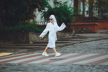 A glamorous lady in a white bathrobe with a white towel on her head and elegant sunglasses is walking along the street of a big city
