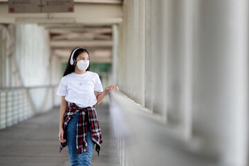 Asian young woman walking on bridge with her headphones while wearing face mask as protection against infectious diseases during coronavirus pandemic. Basic equipment to help keep us safe new life.