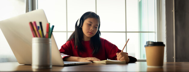 Female student wears headphones sitting at table to learning and note, using laptop for homework during covid19 pandemic and lock down, Social Distance, New life of Student on internet. Banner Size