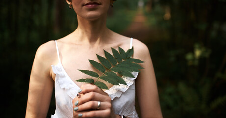 Cropped shot of beautiful tender young bride in romantic white dress posing against green forest background, holding fern leaf at her chest. Unrecognizable female relaxing outdoors among plants