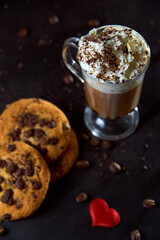 Close up of a glass cup of coffee with whipped cream and chocolate on it, chocolate chip cookies and roasted coffee beans on dark background. Concept of ready to eat food, tasty snack. Selective focus