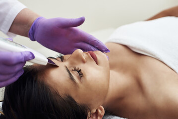 Fototapeta premium A woman makes an ultrasonic cleaning of the face and skin to a client. Modern equipment. Young pretty woman receiving treatments in beauty salons and lying on the couch