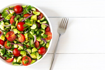 Salad of fresh ripe tomato, onion, avocado, cucumber and parsley and olive oil, salad in a white plate with a fork and towel on a white wooden background