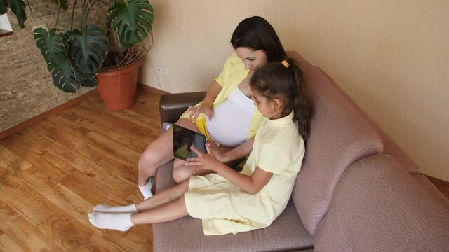 A young pregnant woman with her daughter is watching an ultrasound video on a tablet. Waiting for birth, the appearance of a child. Pregnant girl with a tablet on the couch.