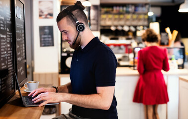 Young man with headphones and laptop indoors in cafe, working.
