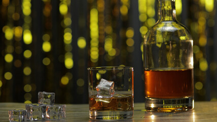 Golden whiskey, cognac or brandy in glass with ice cubes and bottle on table. Shiny background